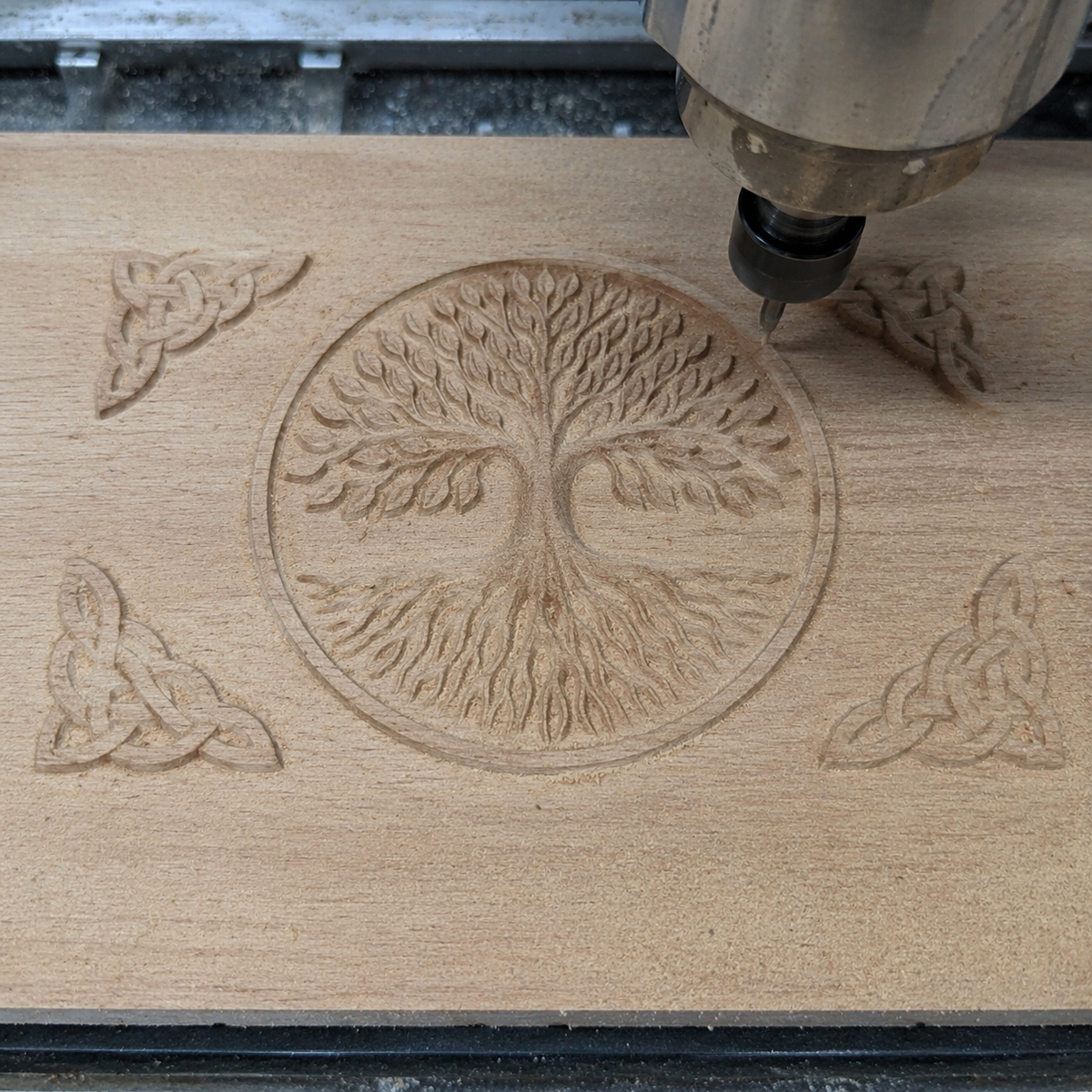 CNC machine carving Celtic tree of life design into wood panel, visible toolpaths forming circular tree and corner knotwork details, partially carved surface showing depth progression and clean cut lines, ideal example of STL file in active CNC carving process